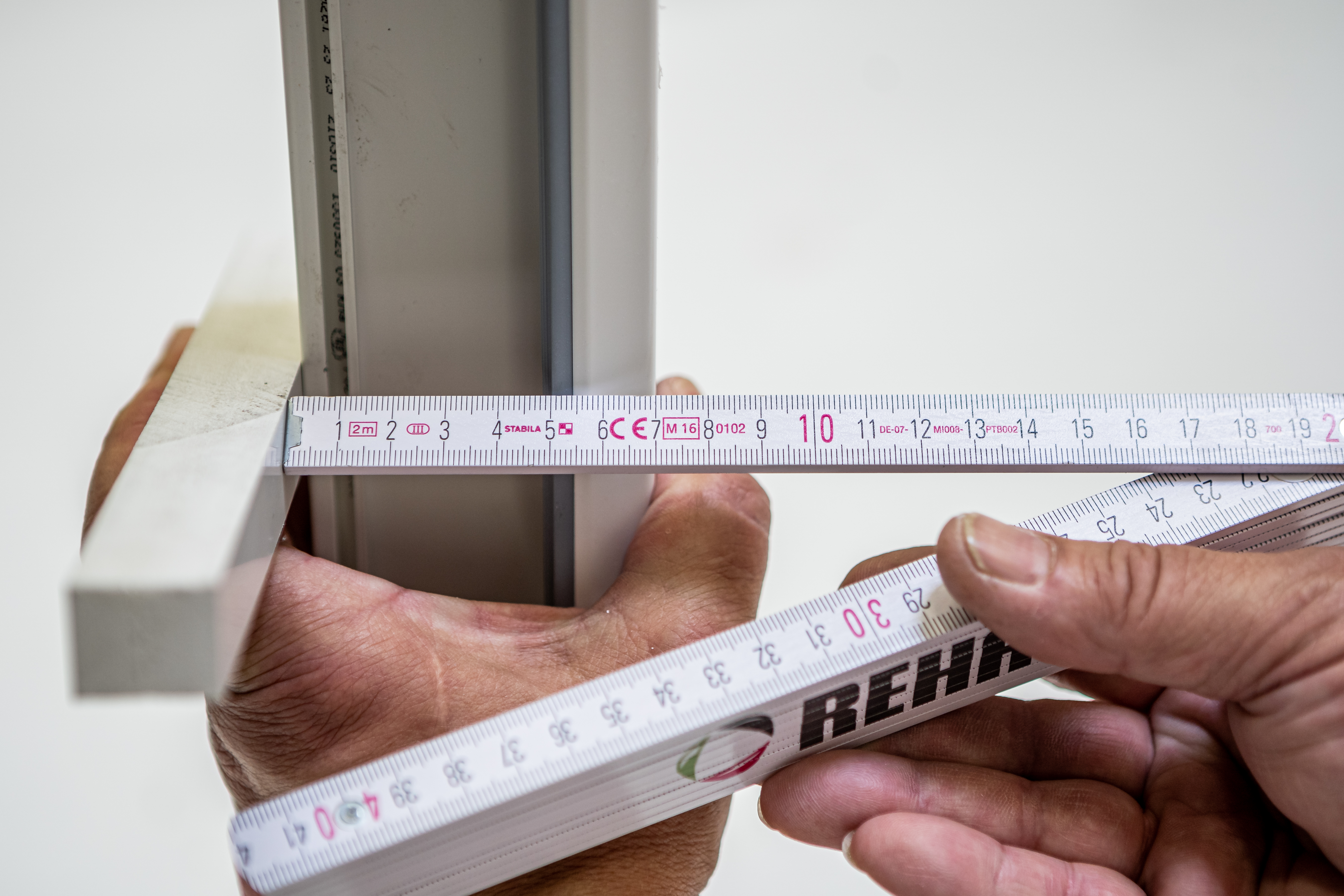 Eine Nahaufnahme von Händen, die einen Zollstock an einem Fensterrahmen anlegen, um die genaue Tiefe oder Breite einer Dichtungsposition zu messen.